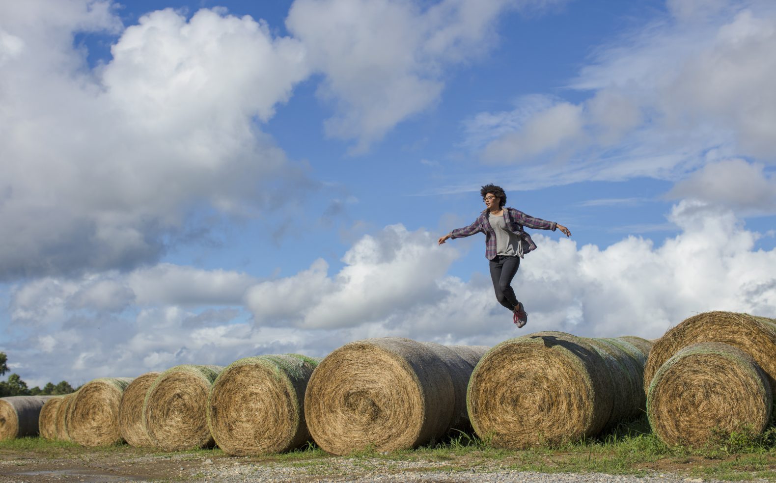 A young woman wearing jeans and a flannel is airborne as she jumps off a row of hay barrels under a blue sky.