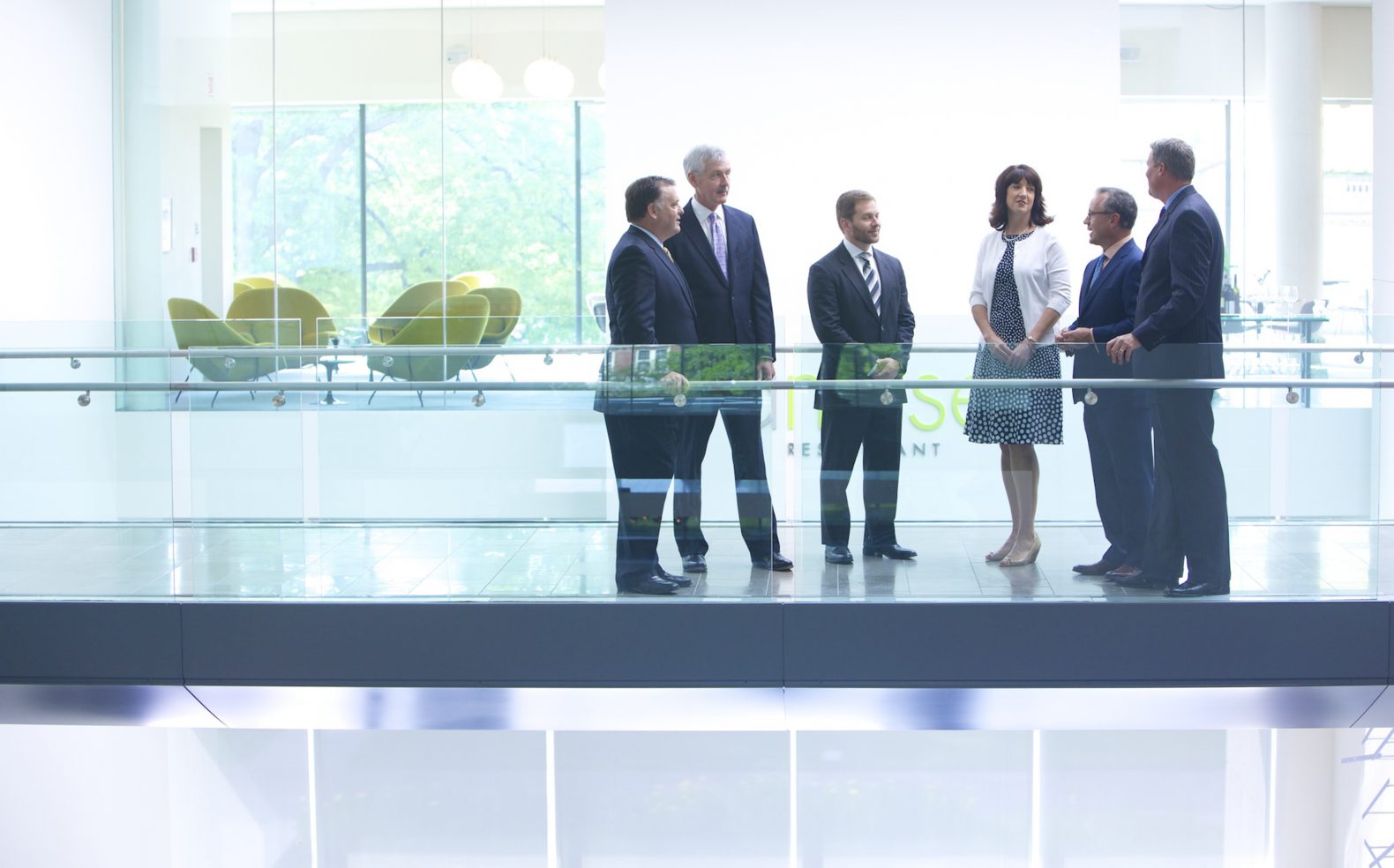 A group of six business professionals in formal business attire congregating in a modern office hallway.