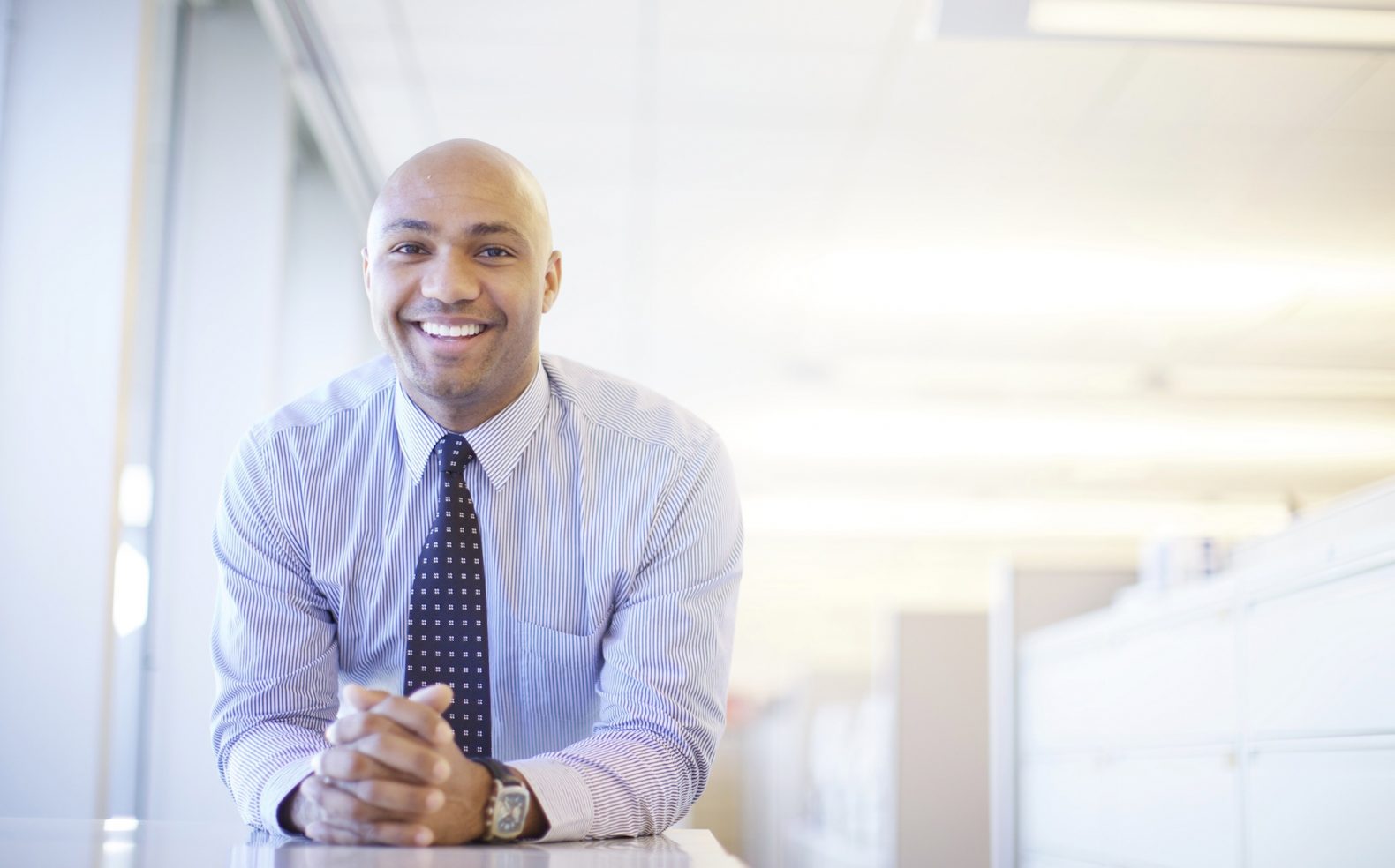 A male professional in business attire leaning on a tall desk with hands clasped, an out of focus office interior in the background.