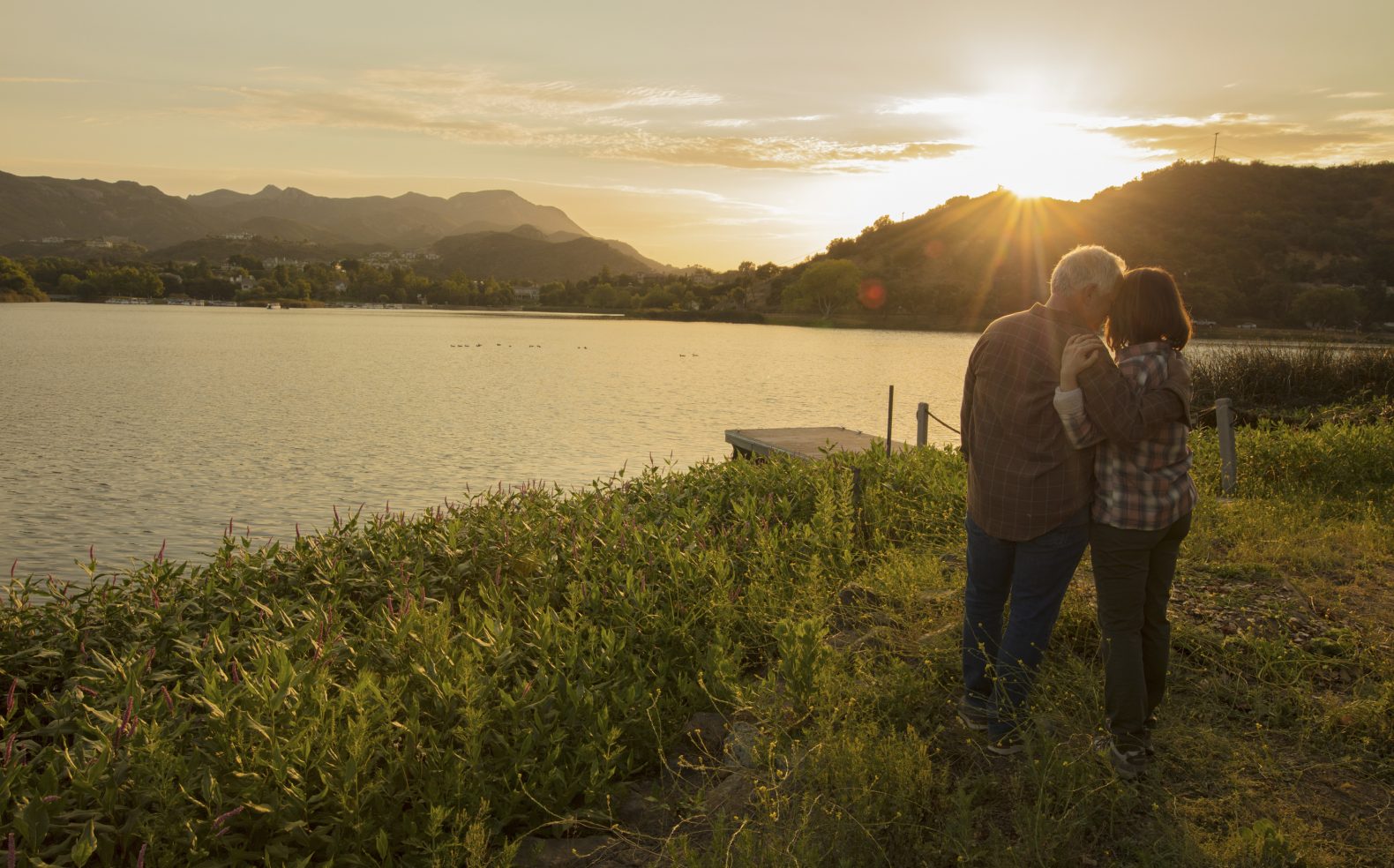 A middle-aged couple standing and embracing in grass next to a dock on a lake while the sun sets over the lake and mountains.