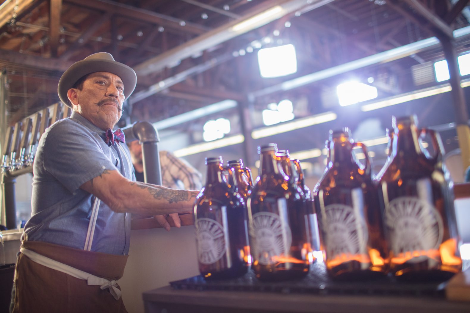 Danny Trejo wearing a fedora at a brewery next to rows of beer growlers.