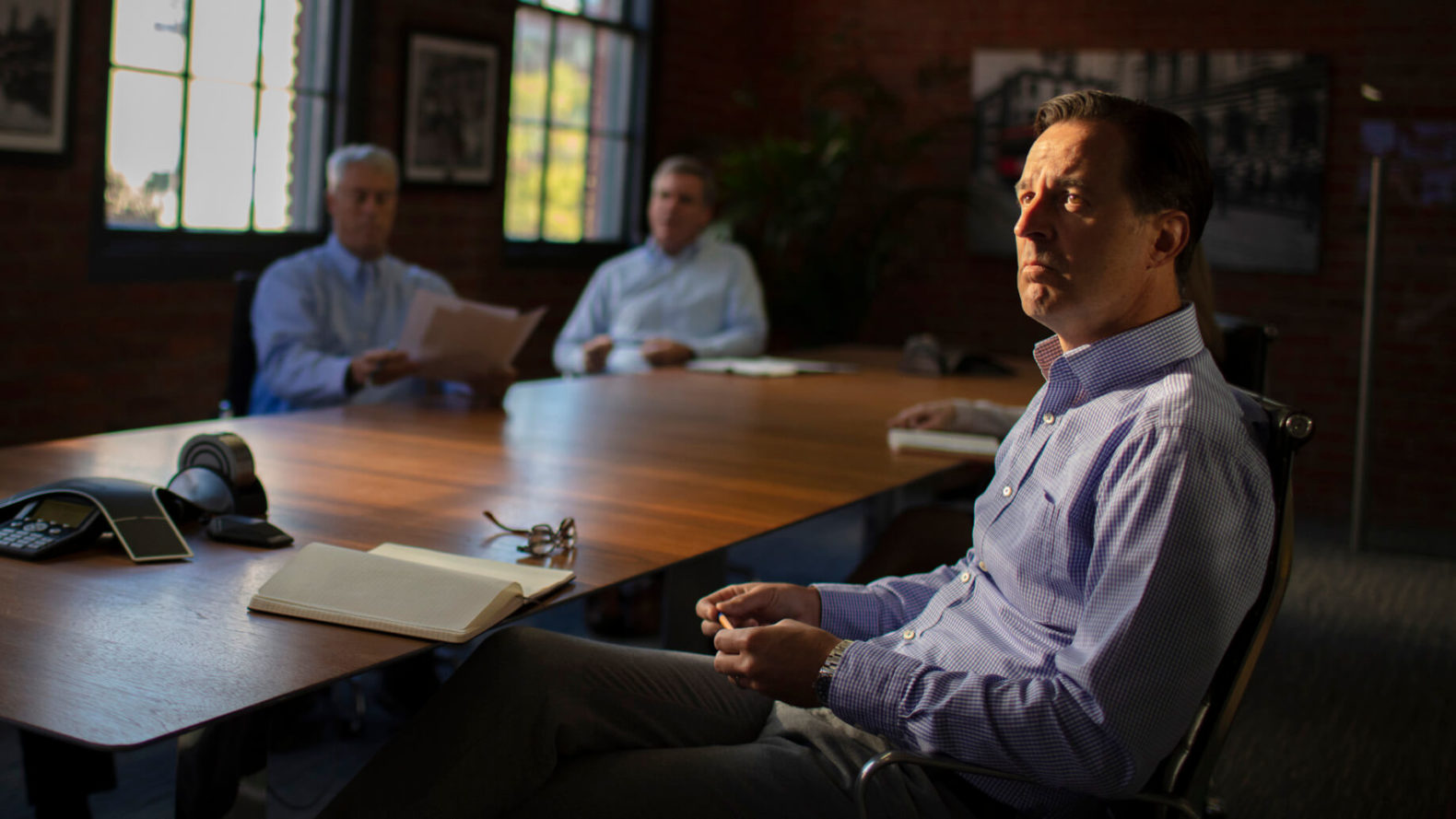 professionals sit at a conference table with notebooks and papers with windows in the background