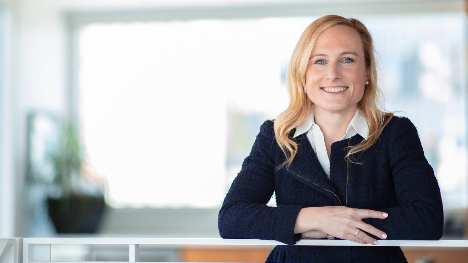 female professional smiles with arms gently crossed on a stair rail with a out of focus office in the background