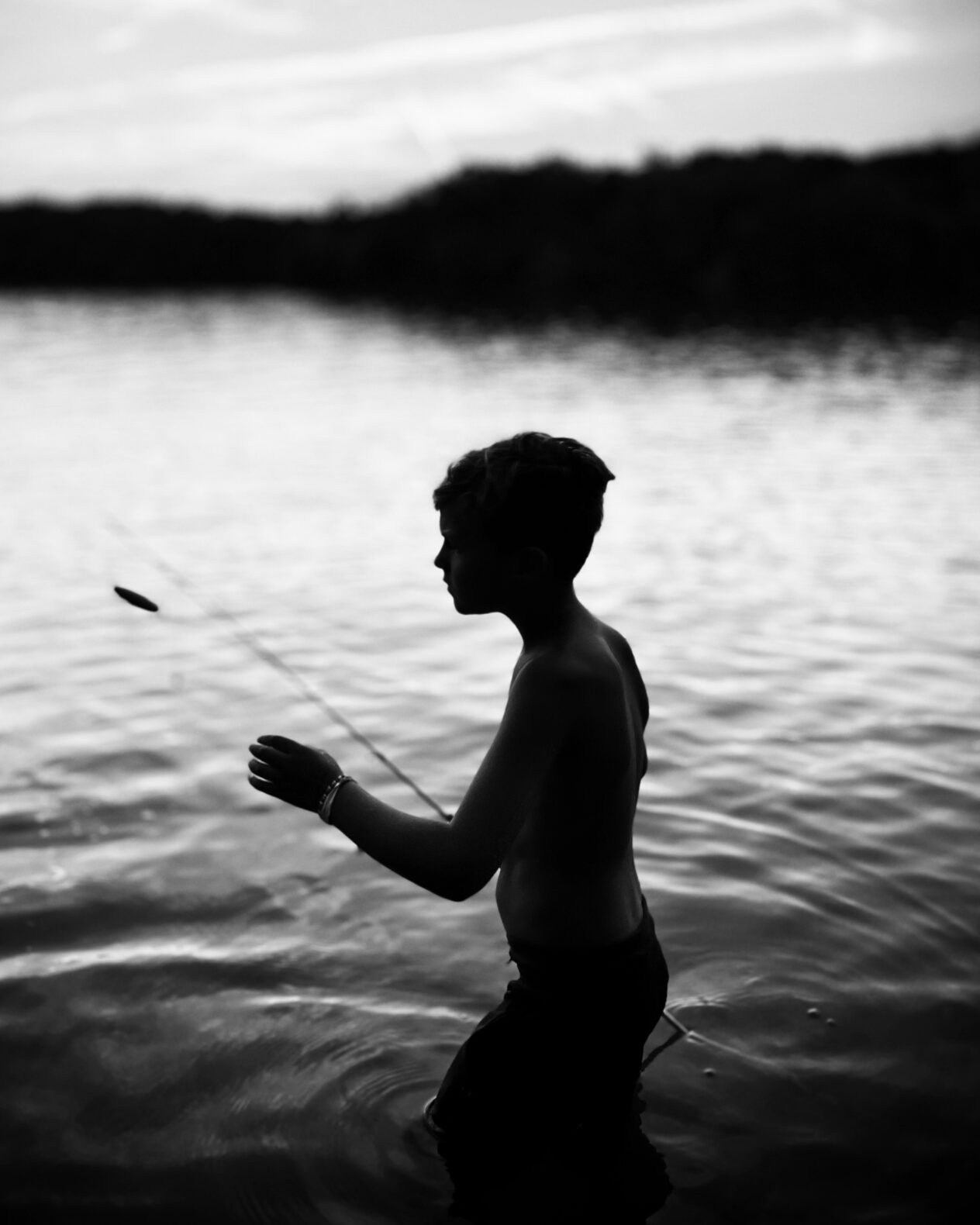 black & white silhouette of boy holding fishing lure standing in lake with tree-lined background