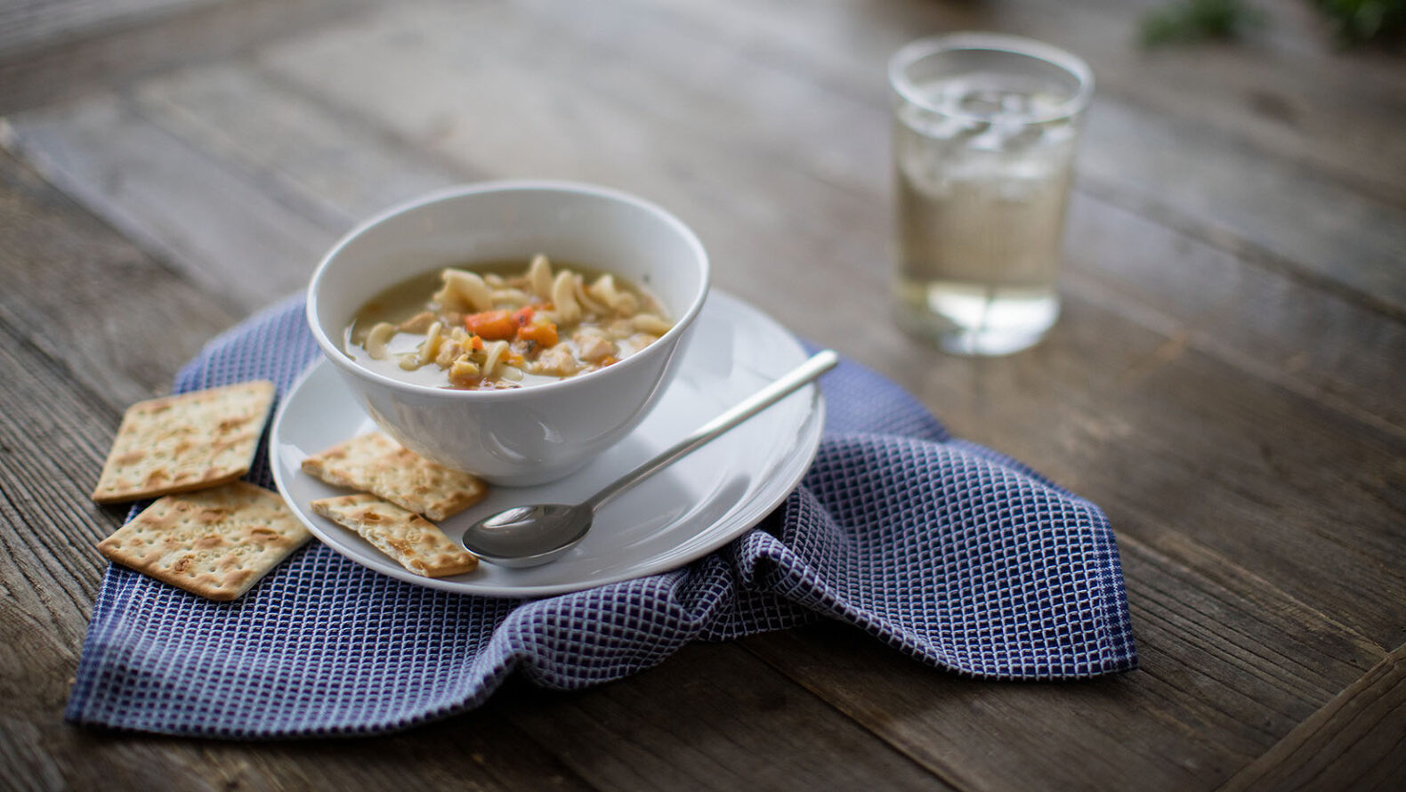 advertising photo of bowl of chicken noodle soup with crackers on a plate with spoon and water cup