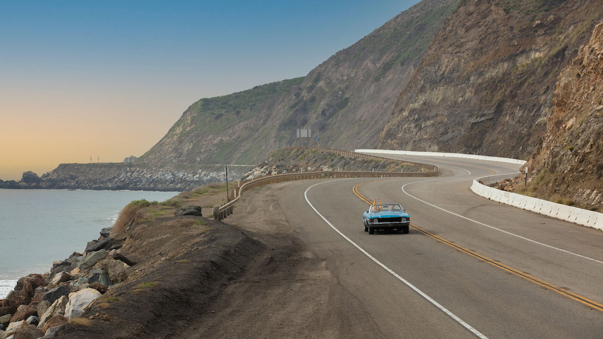 two people driving down coastal highway in blue convertible