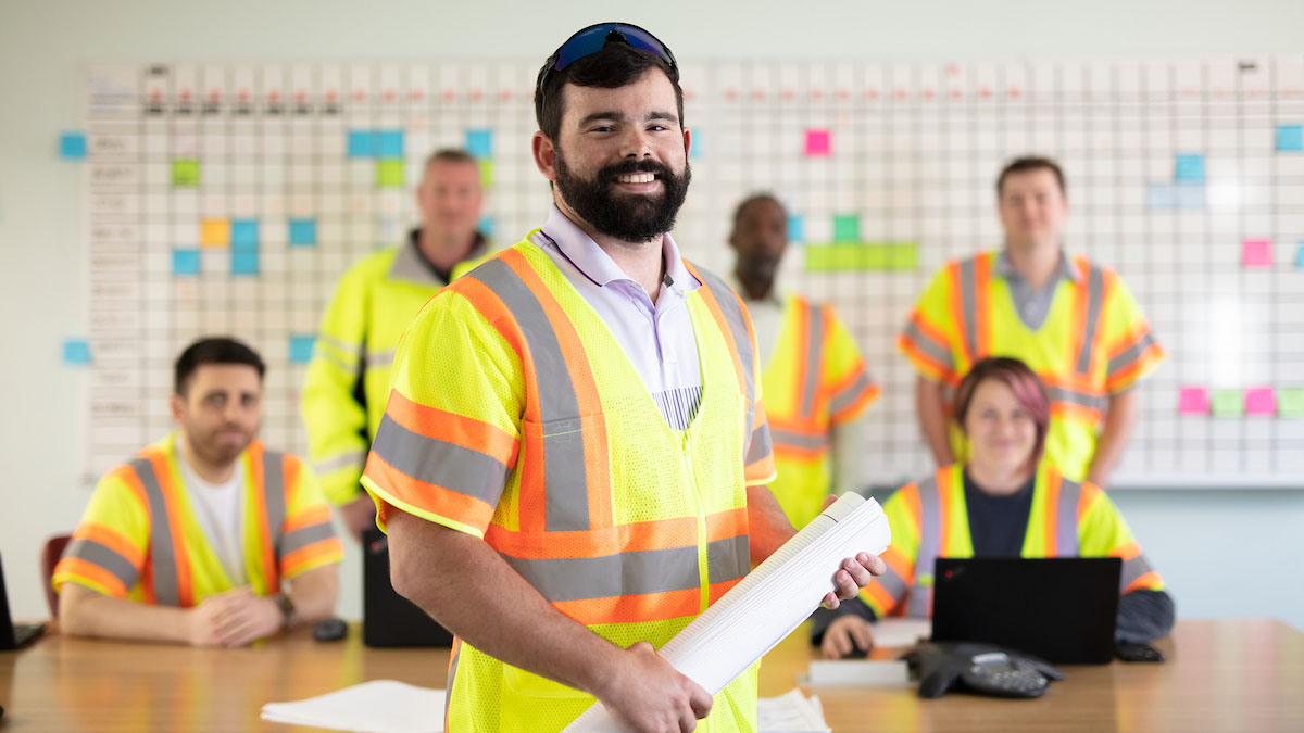 Male employee in highlighter yellow and orange vest with team out of focus in background