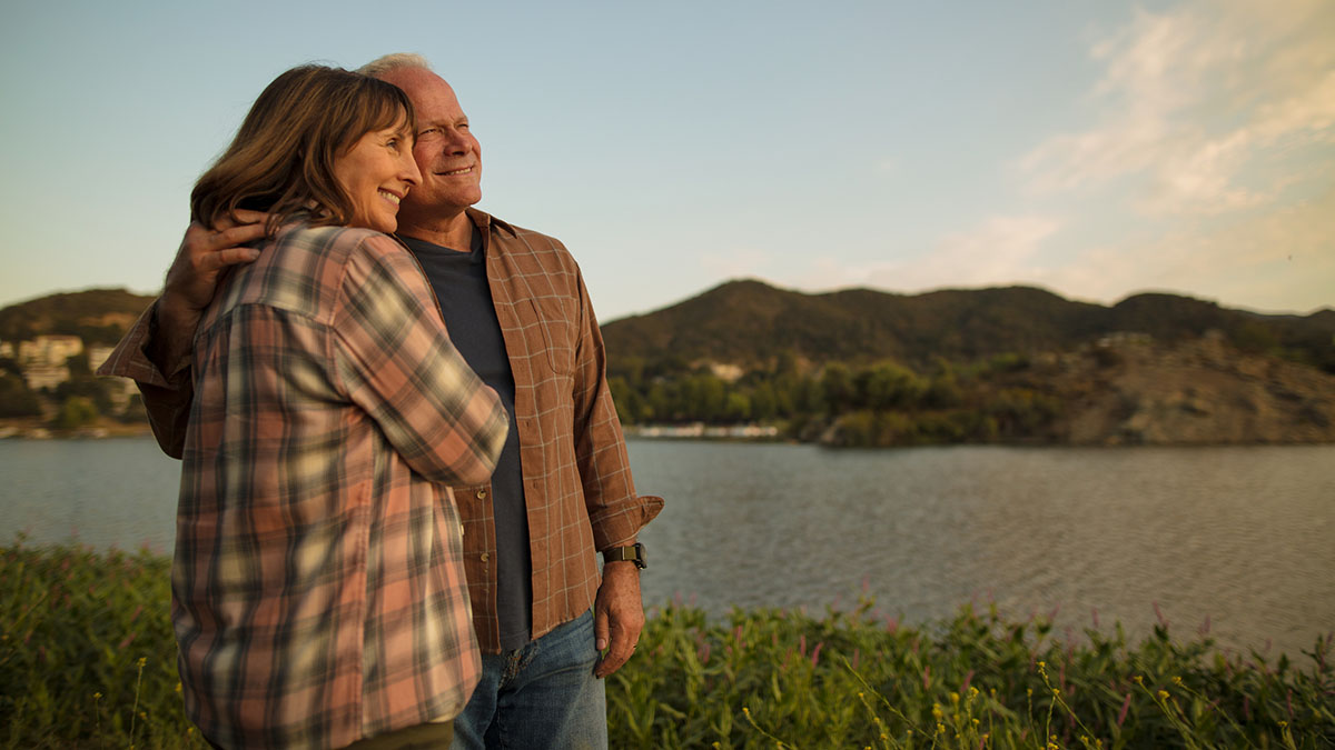 Man and woman in flannel long sleeve shirts stand next to body of water with mountains in background