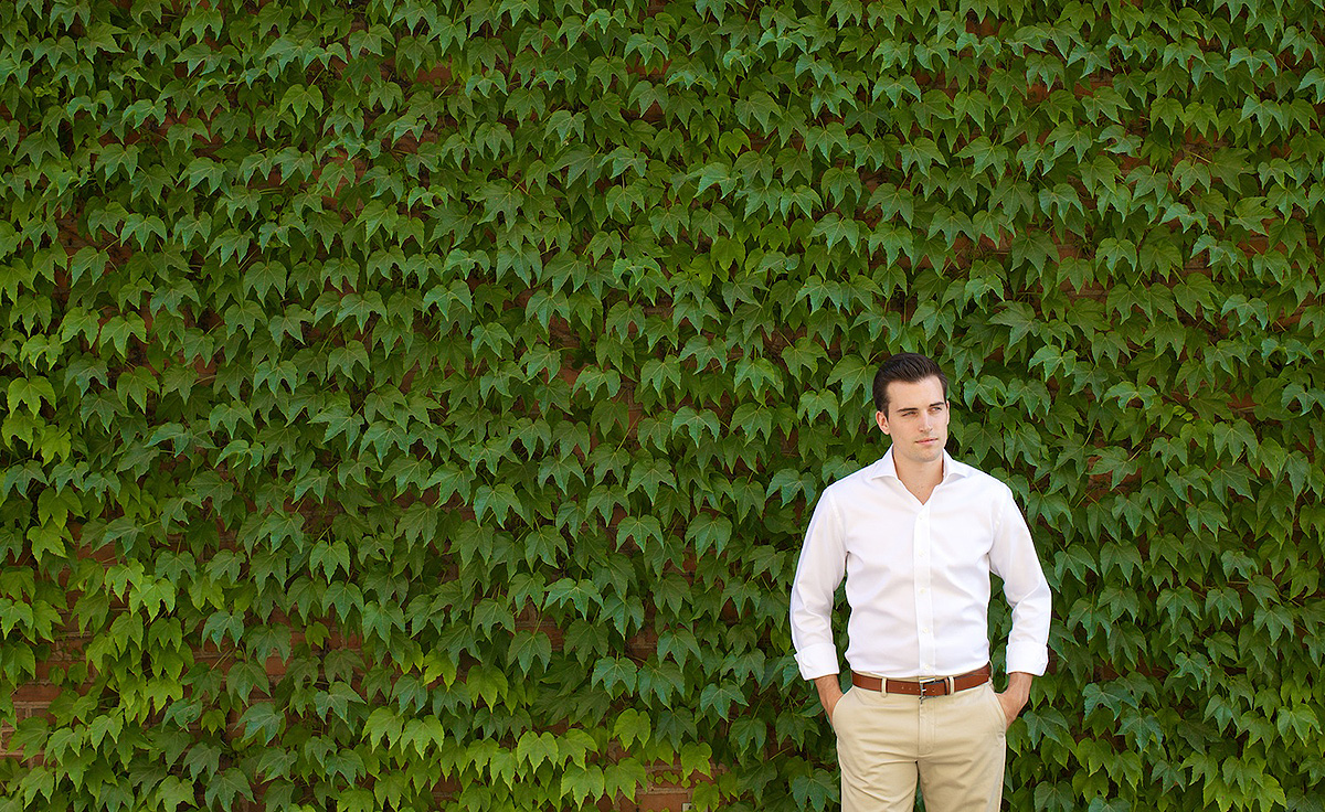 man with slick dark hair in white dress shirt and tan dress pants standing in front of leaf covered wall