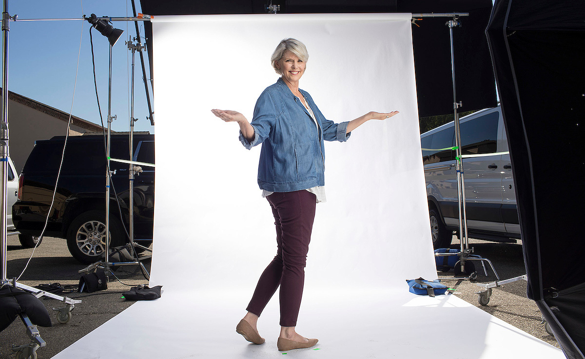 Female model with grey hair in denim jacket and maroon pants standing in front of white screen on photo shoot