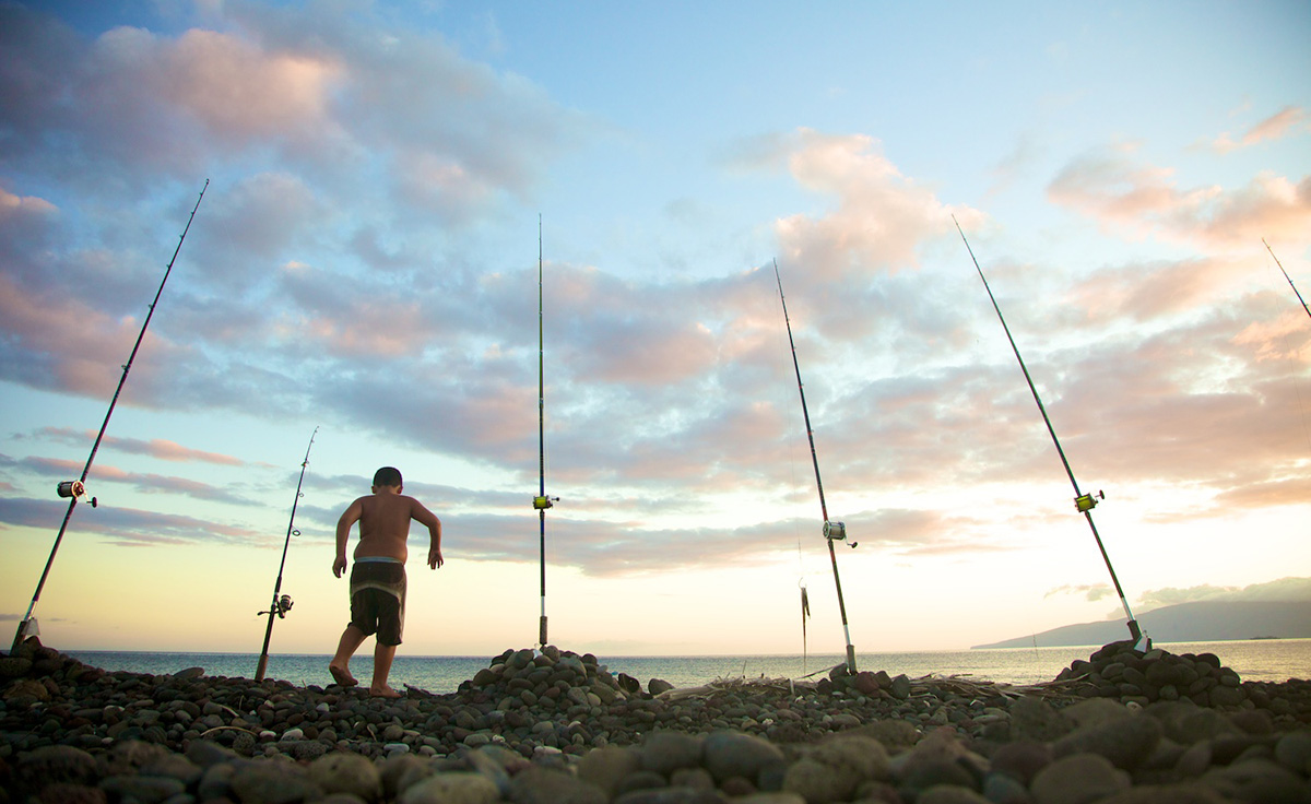 young boy in bathing suit on rocks lined with fishing reels facing open water and mountain