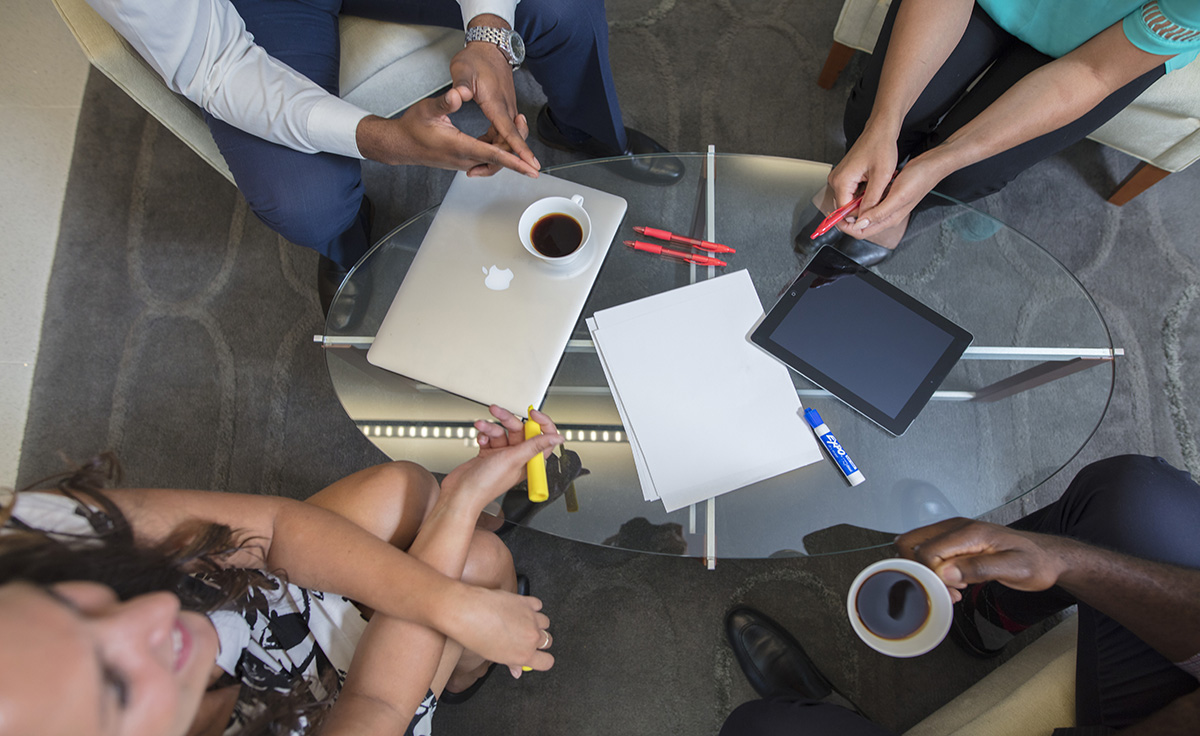 group of employees collaborating at oval see-through table with ipad, macbook, paper, red pens, and markers