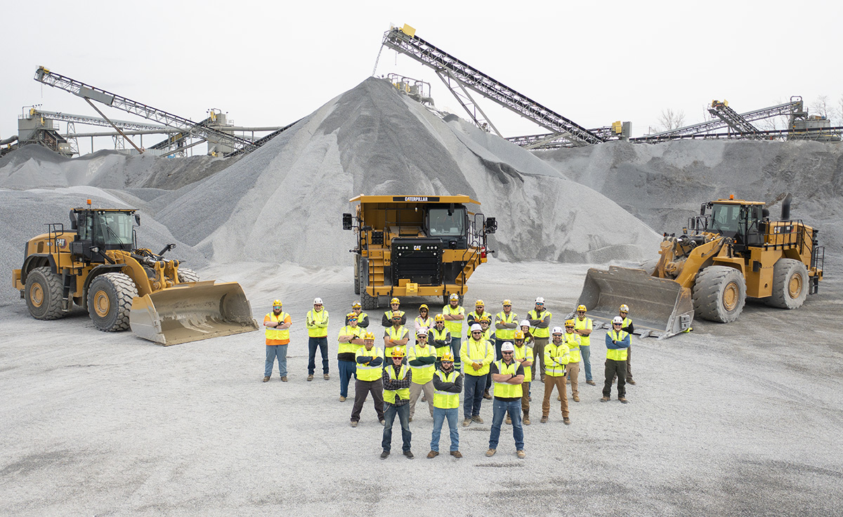 group of construction workers standing in front of piles of stone and CAT vehicles with crossed arms