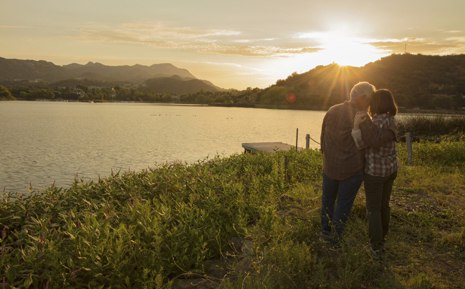 couple embracing in grass looking at dock and lake with mountains in background